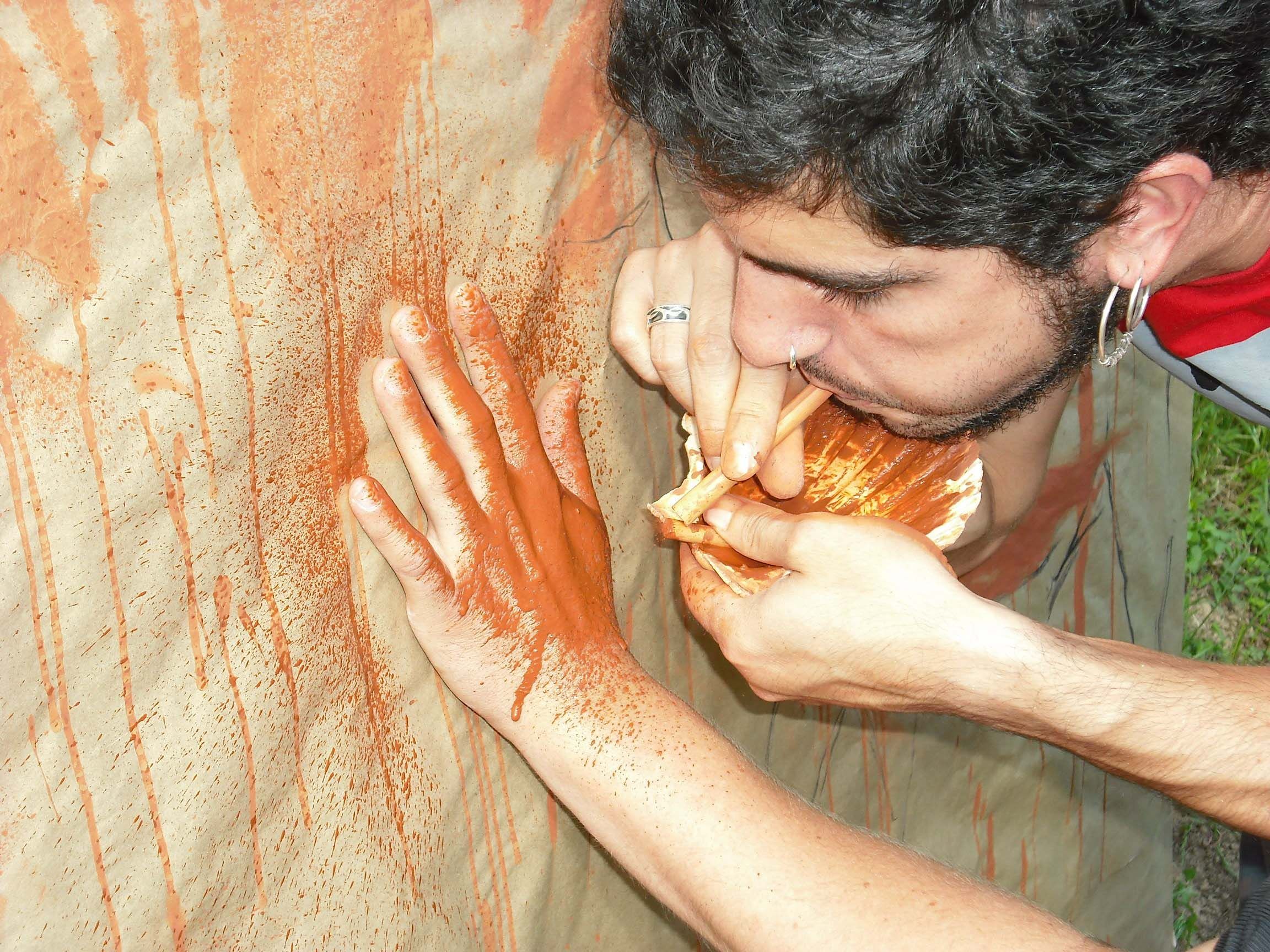 A person blows red paint through a hollow bone onto the canvas on a wall, partially obscured by a hand.