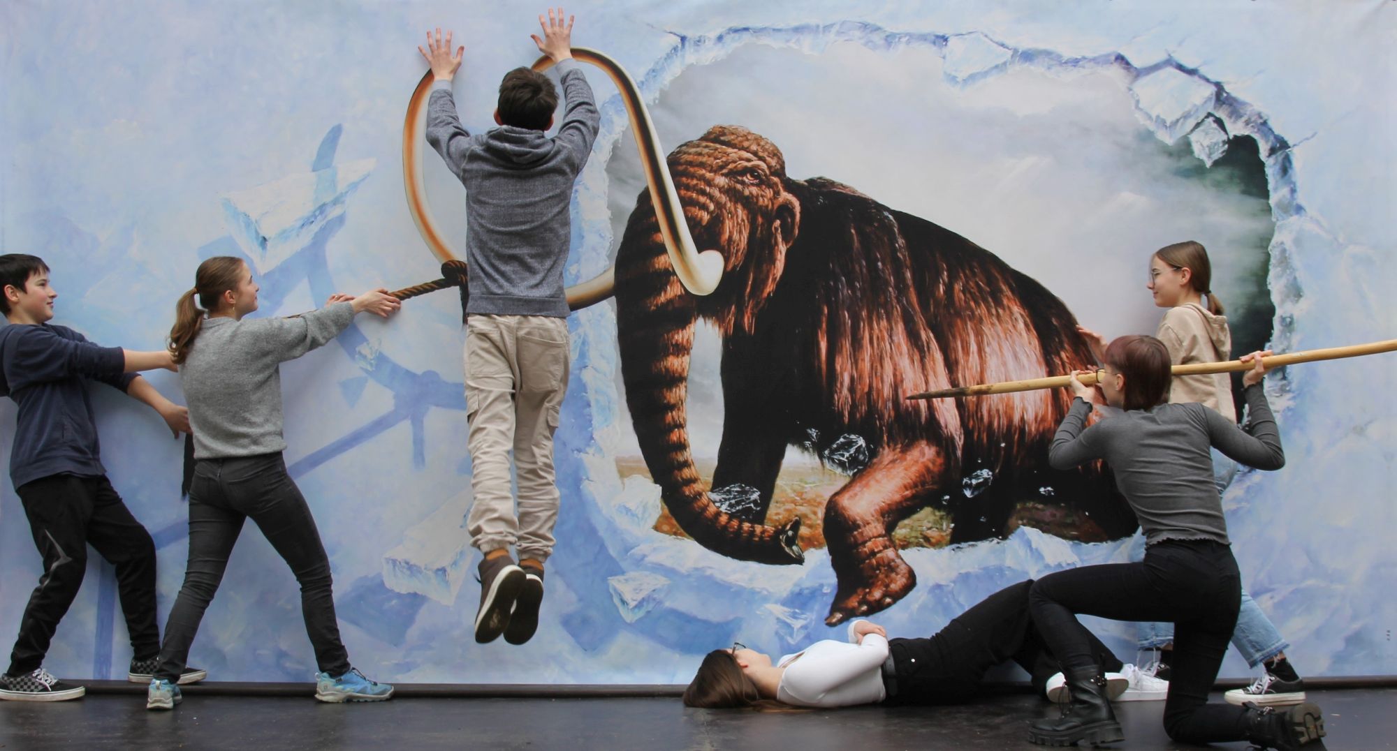 A group poses in front of a large format 3D effect painting on the subject of mammoth hunting