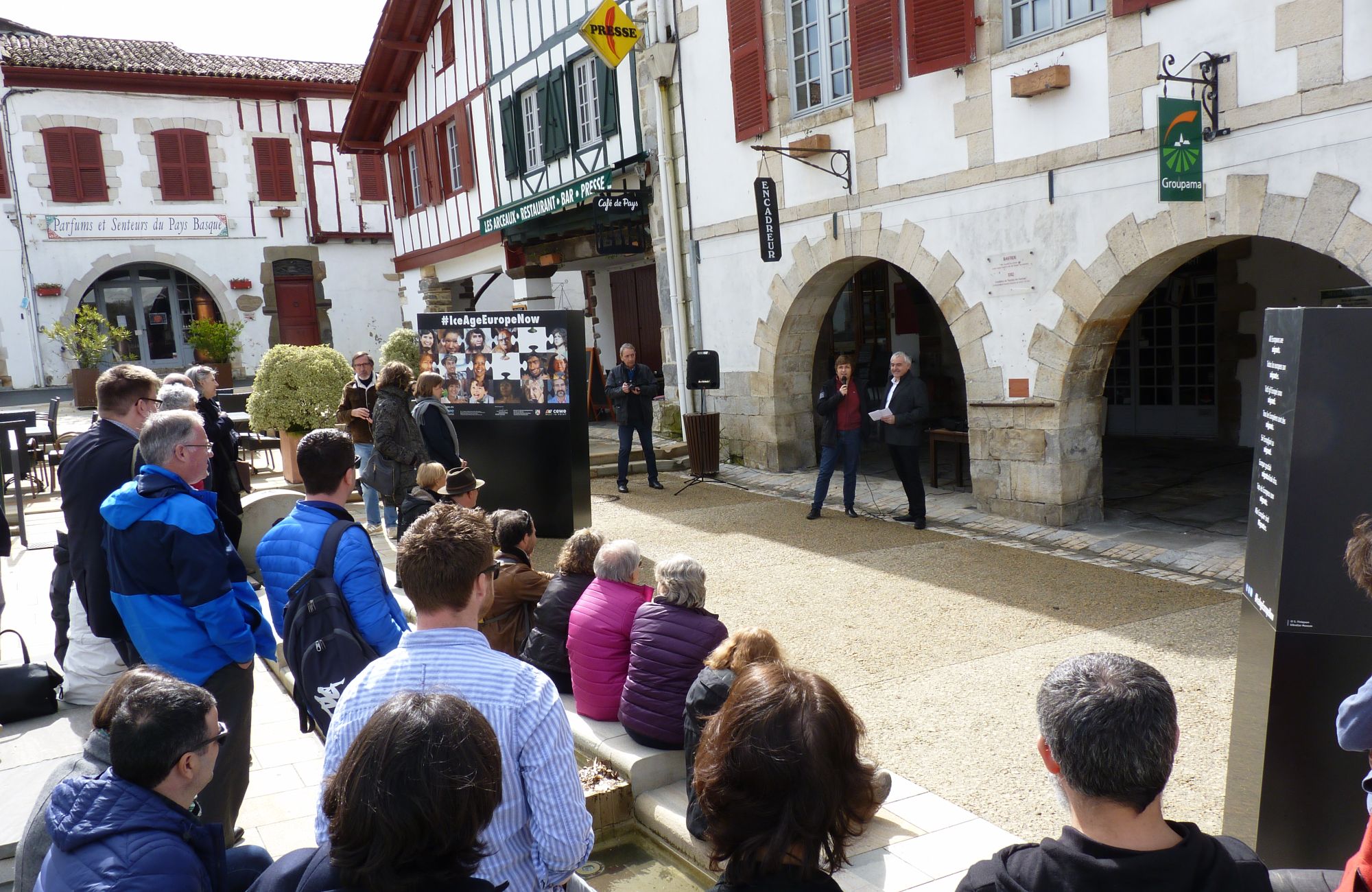 People listening to an opening speech amidst large photo panels in a picturesque town square